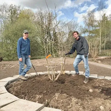 Two volunteers stand on either side of a freshly planted tree in a circular raised bed built with stacks of concrete slabs in Lochhead Gardens