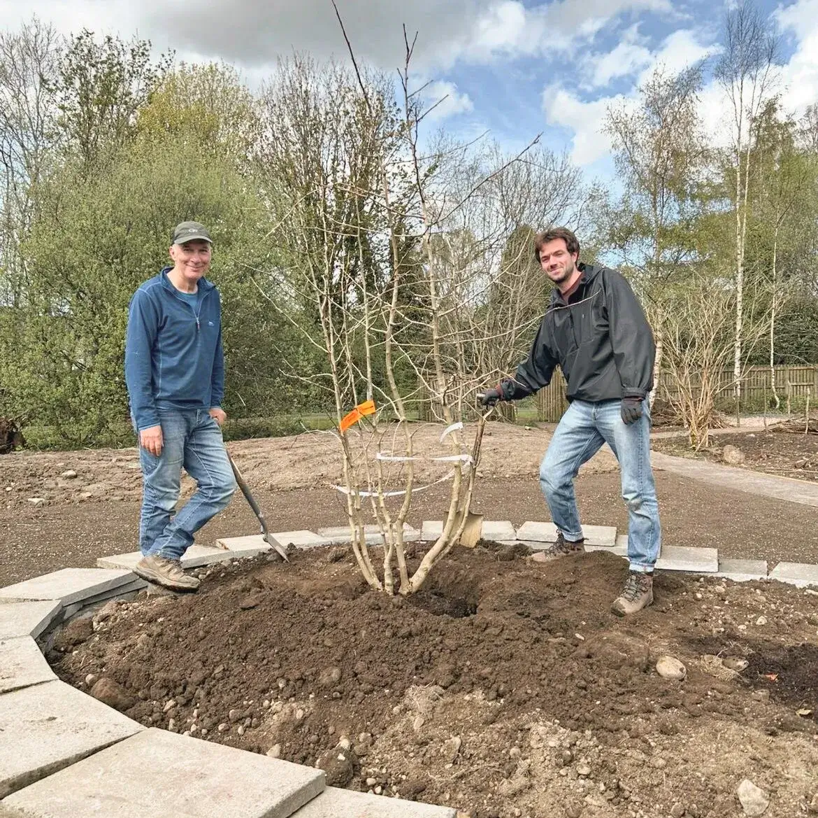 Two volunteers stand on either side of a freshly planted tree in a circular raised bed built with stacks of concrete slabs in Lochhead Gardens