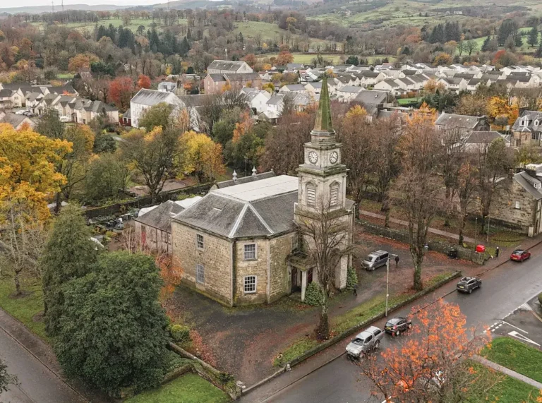 High-angle aerial view of Lochwinnoch Parish Church, showing its stone octagonal building and tall steeple surrounded by trees and village houses.