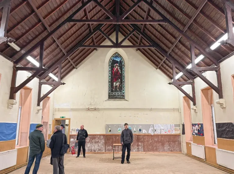 Interior view of the spacious Guild Hall featuring a high, vaulted wooden timber ceiling, a stained glass window on the far wall, and a group of people standing on the open floor.
