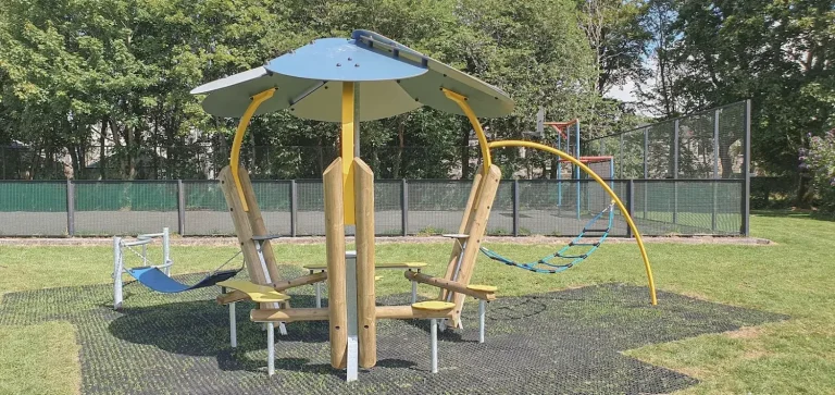An outdoor modern park shelter for teenagers, featuring a blue circular canopy roof, timber and curved steel pillars with integrated seating, and attached hammock-style swings on a rubber mat floor.